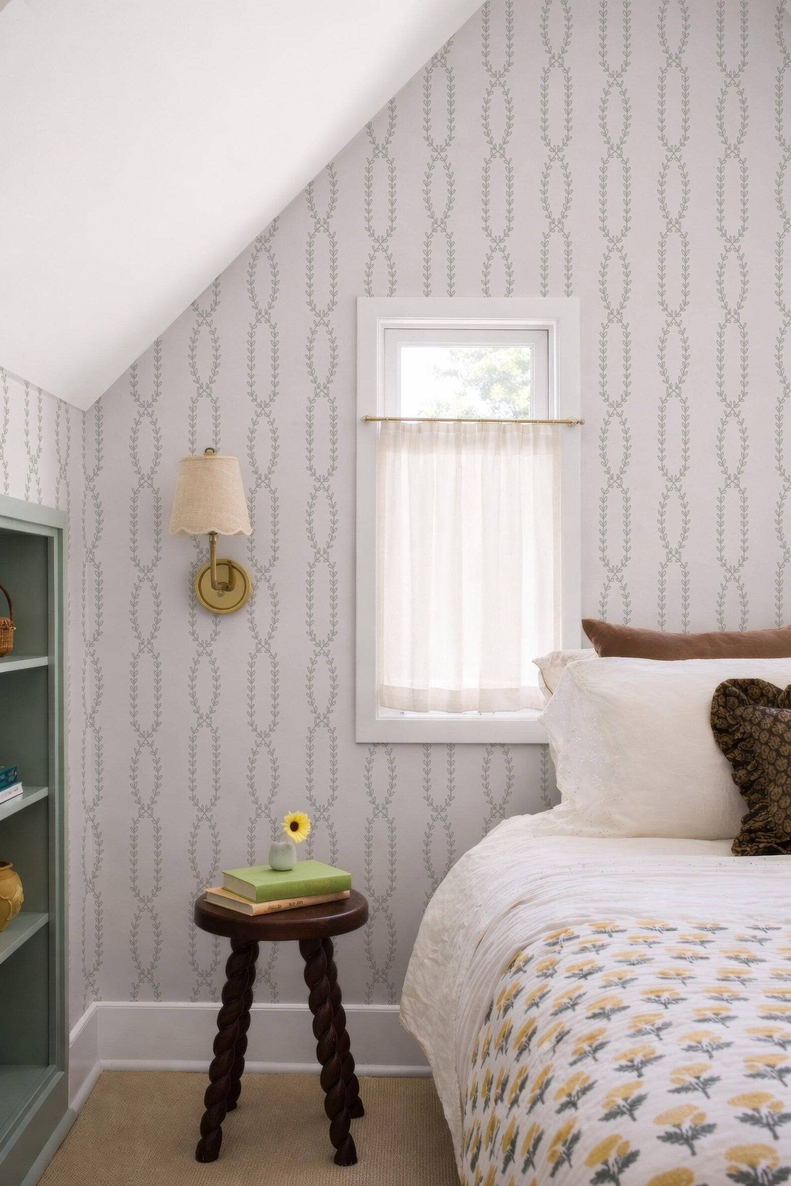 a cozy bedroom with a bed, a small wooden table, and a window with a white curtain. The room has a neutral color scheme with gray and white patterned wallpaper, and the bed is adorned with a white and yellow floral comforter.