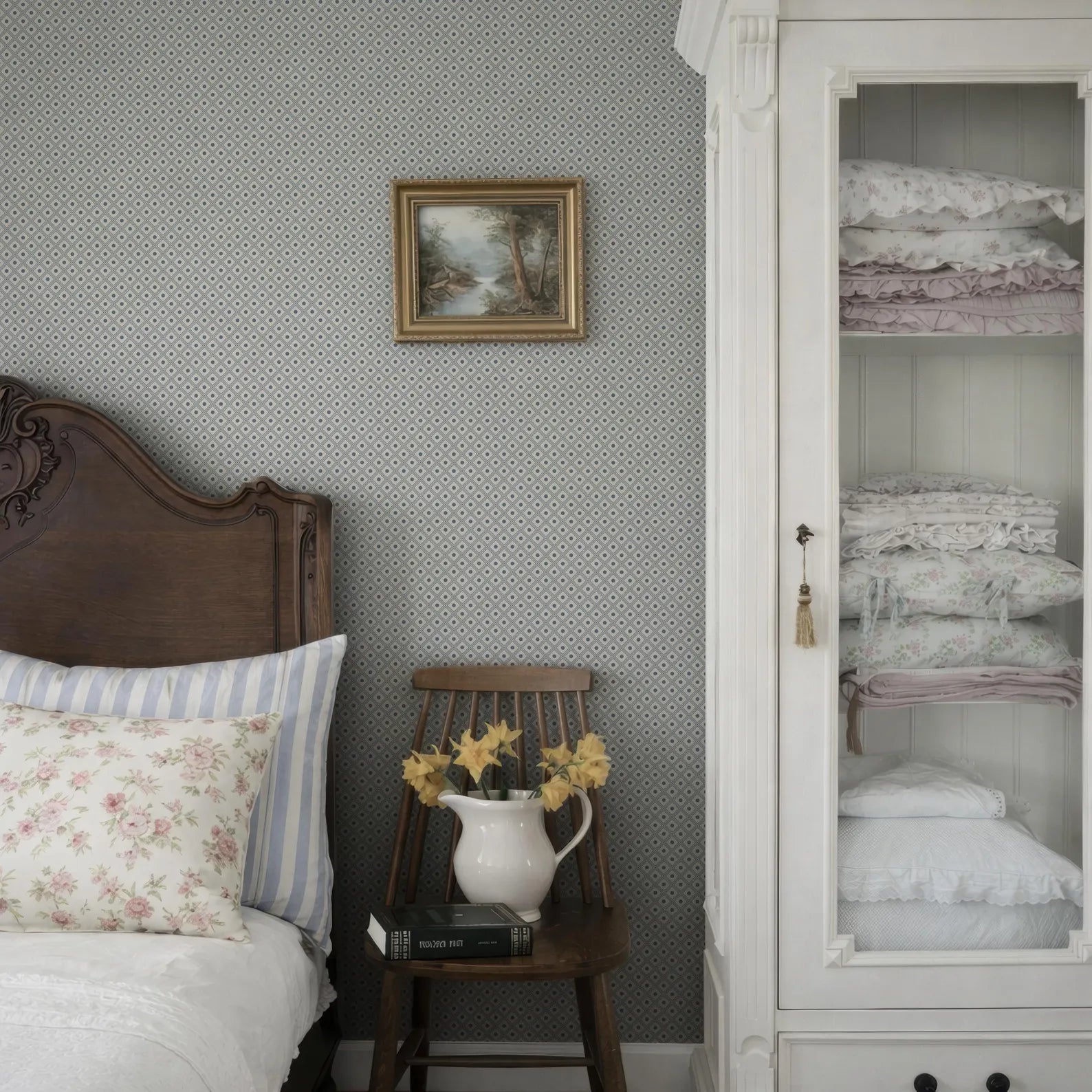 a cozy bedroom scene with a wooden headboard, a white bed, a wooden chair with a vase of yellow flowers, and a white bookshelf filled with books and blankets.