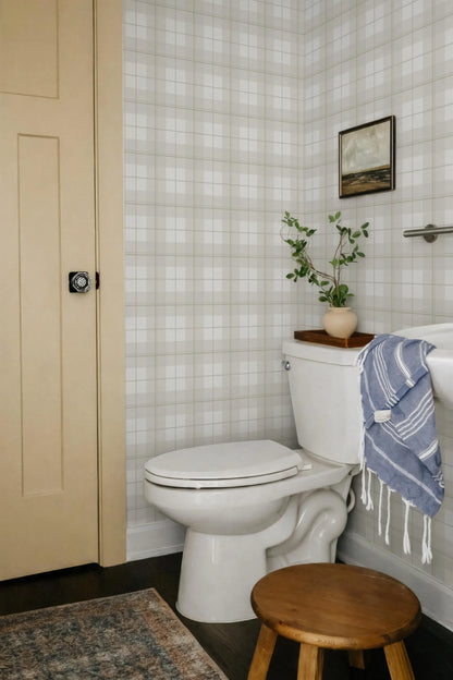 a bathroom with a white toilet, a wooden stool, and a blue and white striped towel hanging on a rack. The walls are covered in a checkered pattern, and there is a painting above the toilet.