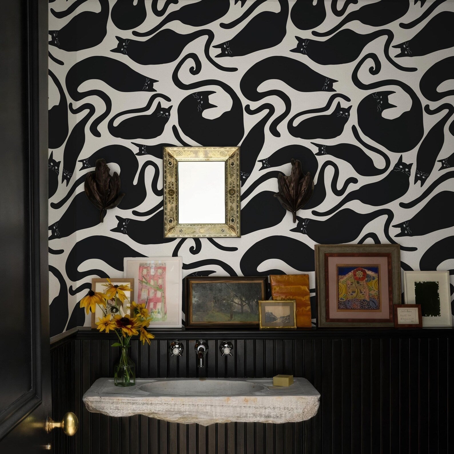 a bathroom with a black and white cat wallpaper, a white sink, and a shelf with framed pictures and a vase of sunflowers.