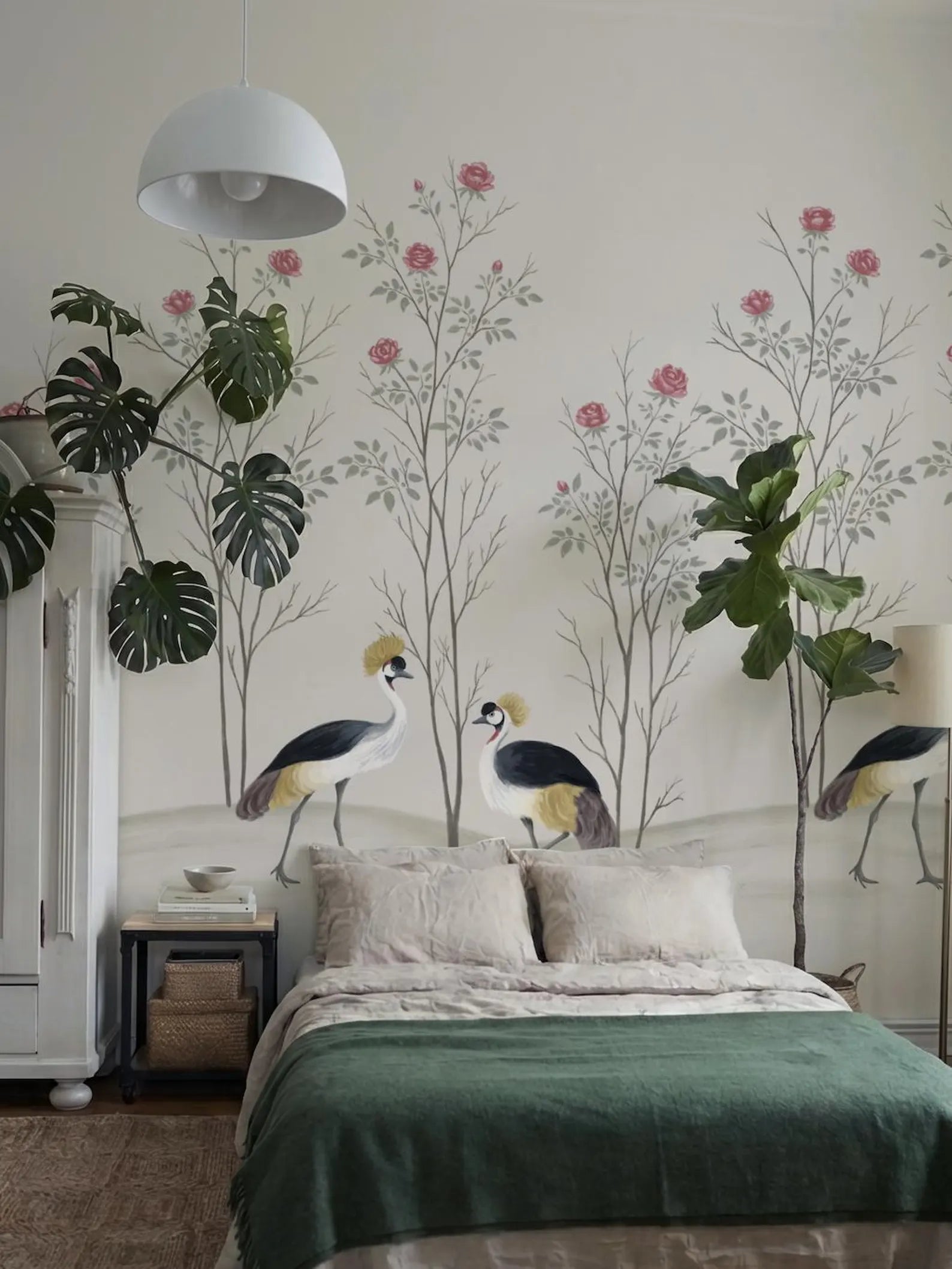 a bedroom with a large mural of a forest scene on the wall, including two birds and pink flowers. The room has a green bedspread, a white lamp, and a white dresser.