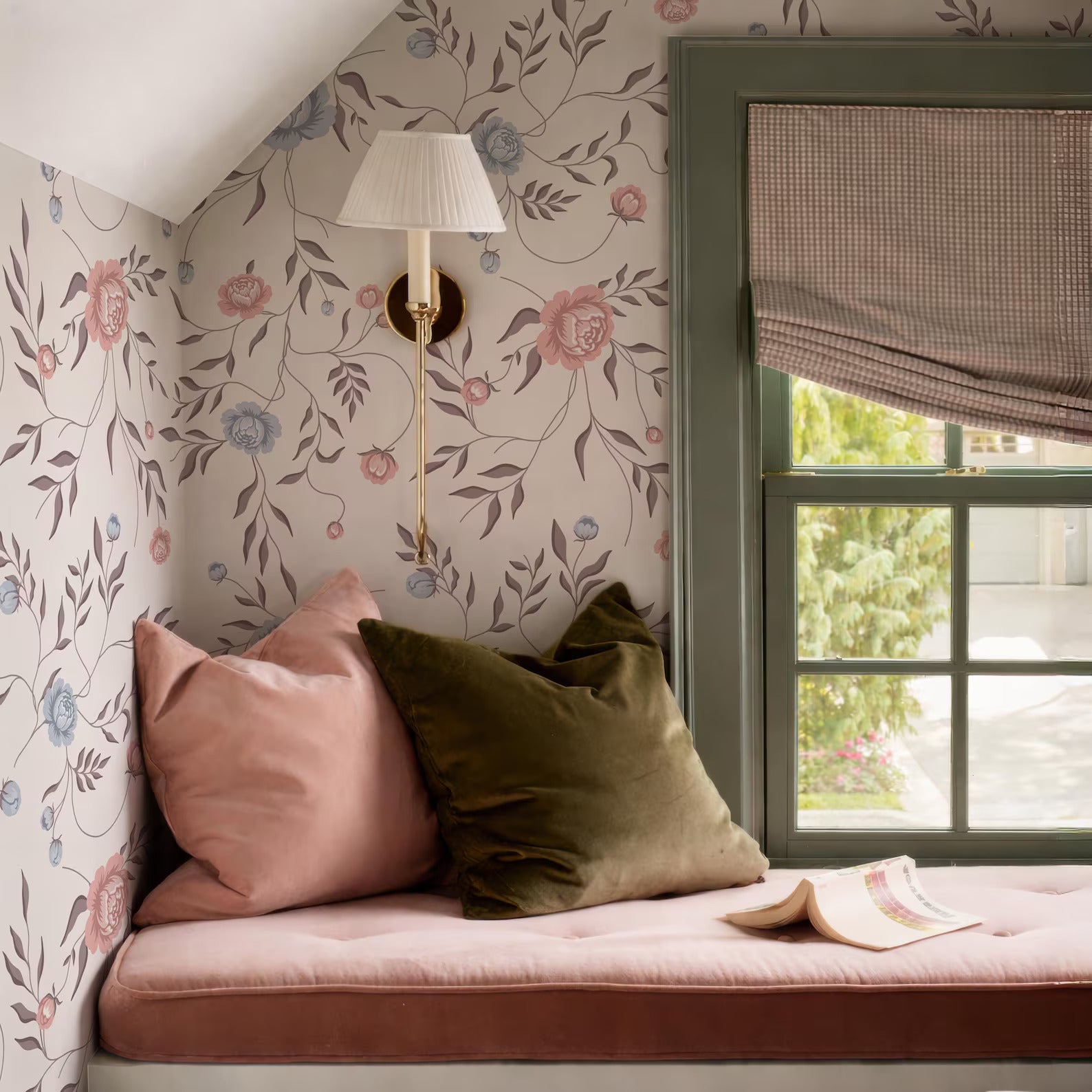 a cozy corner of a room with a floral wallpaper, a window with a green frame, and a lamp on the wall. There are two pillows on the bed, one pink and one green, and an open book is resting on the bed.