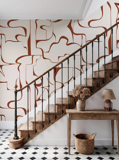 a wooden staircase with a black wrought iron railing, leading up to a white wall with abstract orange and brown designs. A wooden console table with a lamp and a vase of flowers is situated below the staircase, and a woven basket is placed on the floor next to the table.