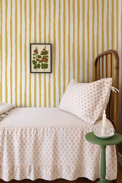 a cozy bedroom scene with a bed, a wooden headboard, and a green side table. The bed is covered in a white and pink polka dot bedspread, and there is a framed picture of flowers hanging on the wall above the bed. The room has a warm and inviting atmosphere, with the yellow and white striped wallpaper adding a touch of color and pattern to the space.