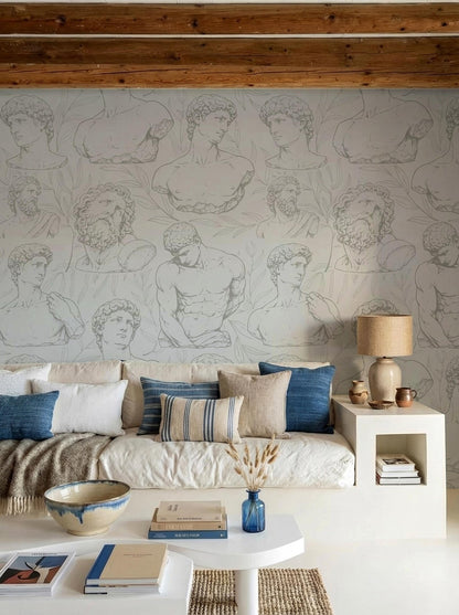 a living room with a white sofa adorned with blue and white pillows, a coffee table with books and a vase of flowers, and a wallpapered wall with various artistic drawings of men's bodies.