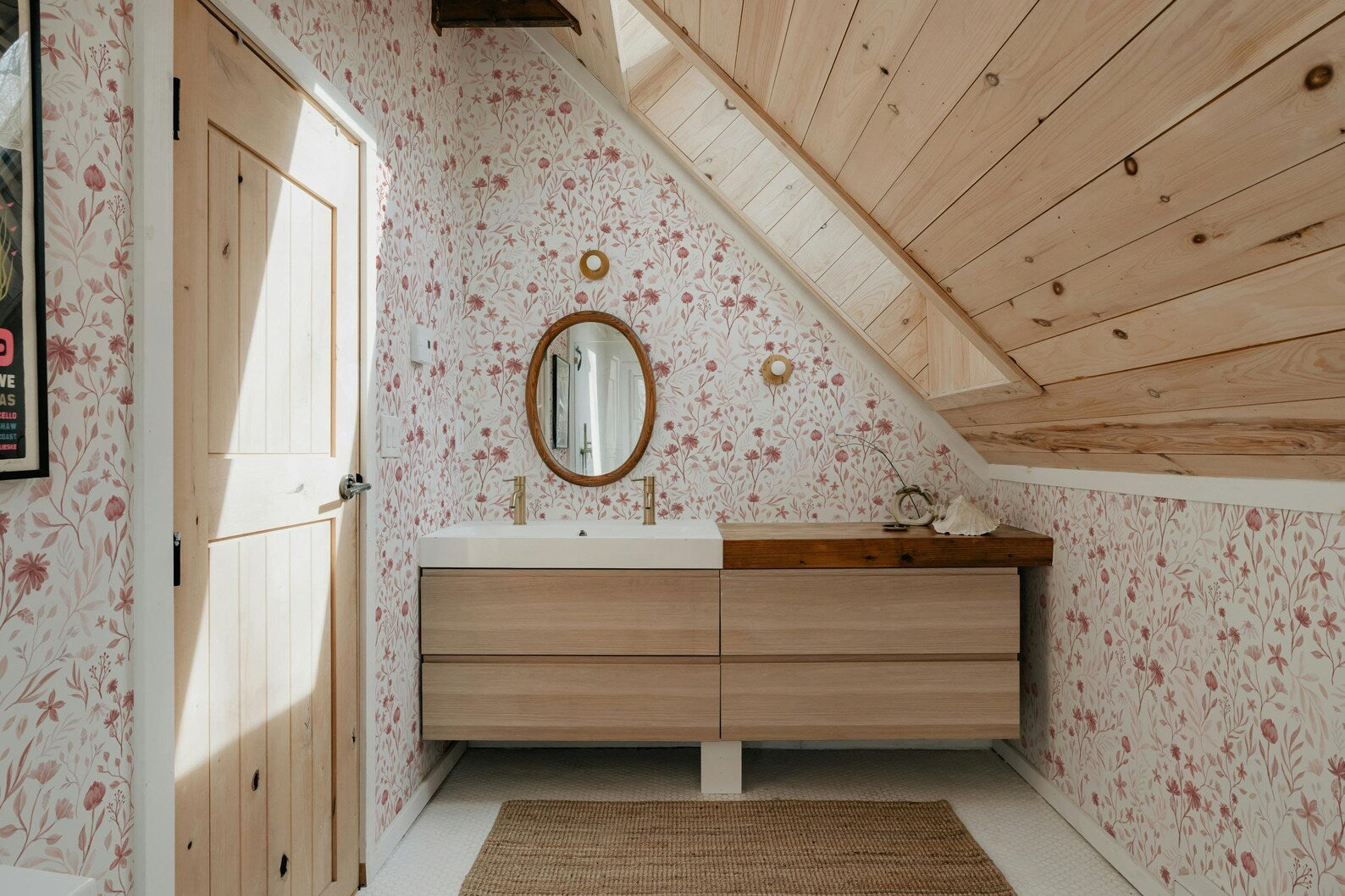a bathroom with a wooden vanity and a mirror above it. The walls are covered in a floral pattern, and the ceiling has a slanted design. The room has a warm and cozy atmosphere, with a rug on the floor and a door on the left side.