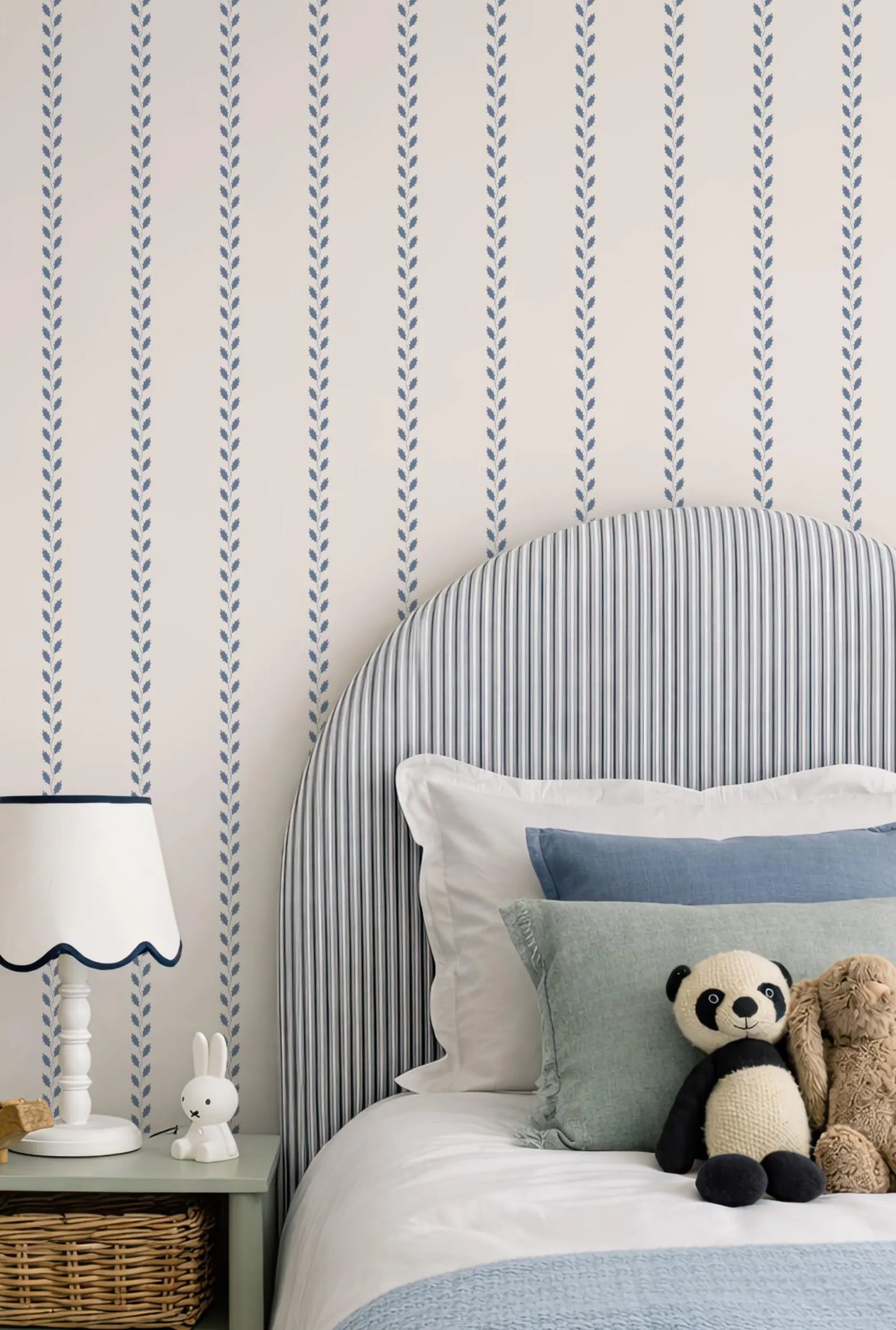 a cozy bedroom scene with a bed, a nightstand, and a stuffed panda bear. The bed has a blue and white striped headboard and is adorned with several pillows. A white lamp is placed on the nightstand next to the bed. A stuffed panda bear is sitting on the bed, adding a playful touch to the scene.