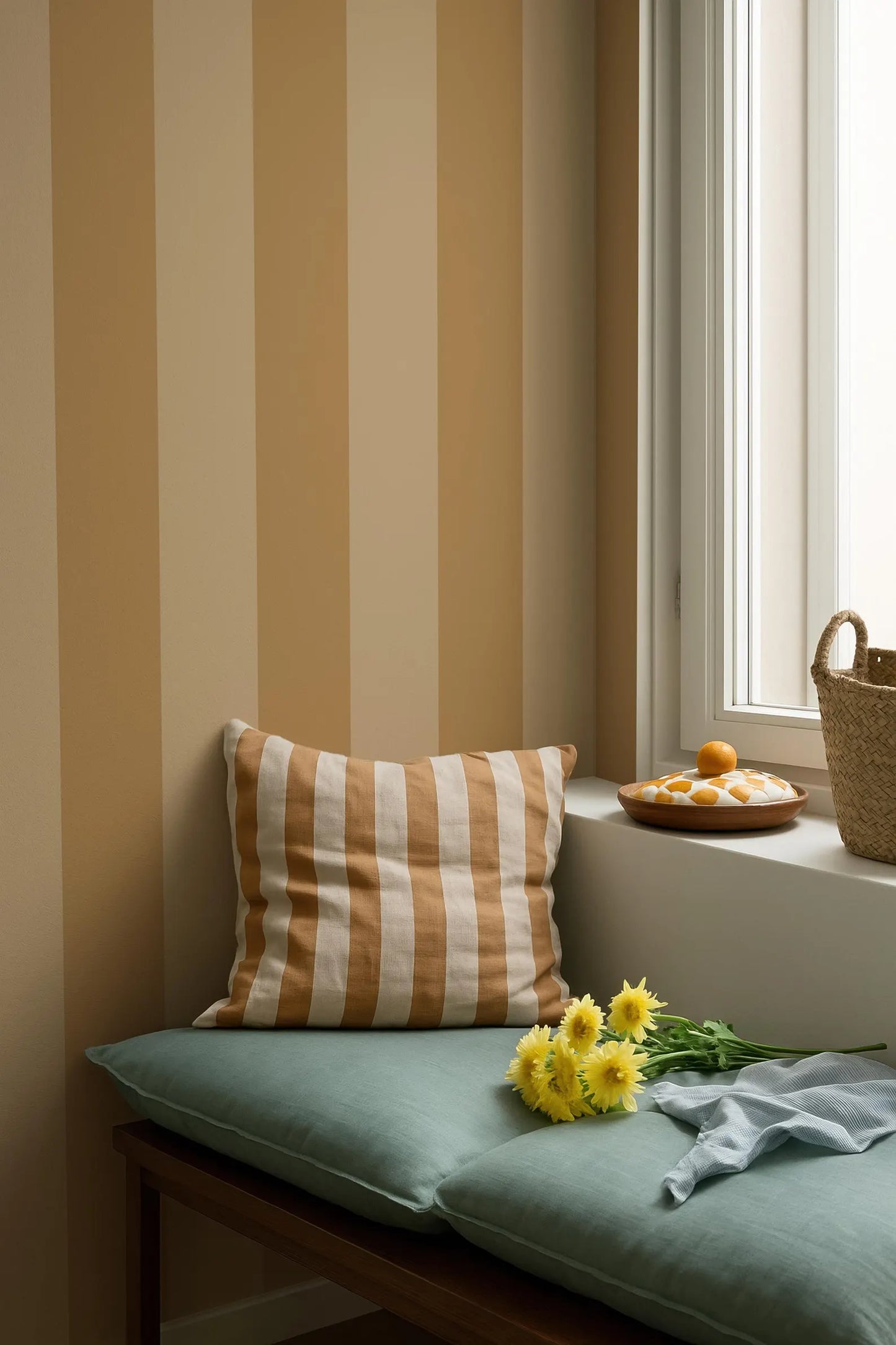 a cozy corner of a room with a striped wall, a green bench, a striped pillow, and a basket of fruit on a windowsill.