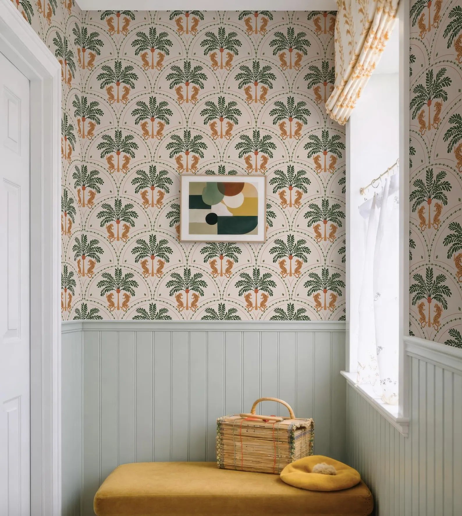 a cozy corner of a room with a yellow bench, a woven basket, and a framed abstract artwork on the wall. The room has a tropical theme, with a patterned wallpaper and a window with white curtains.