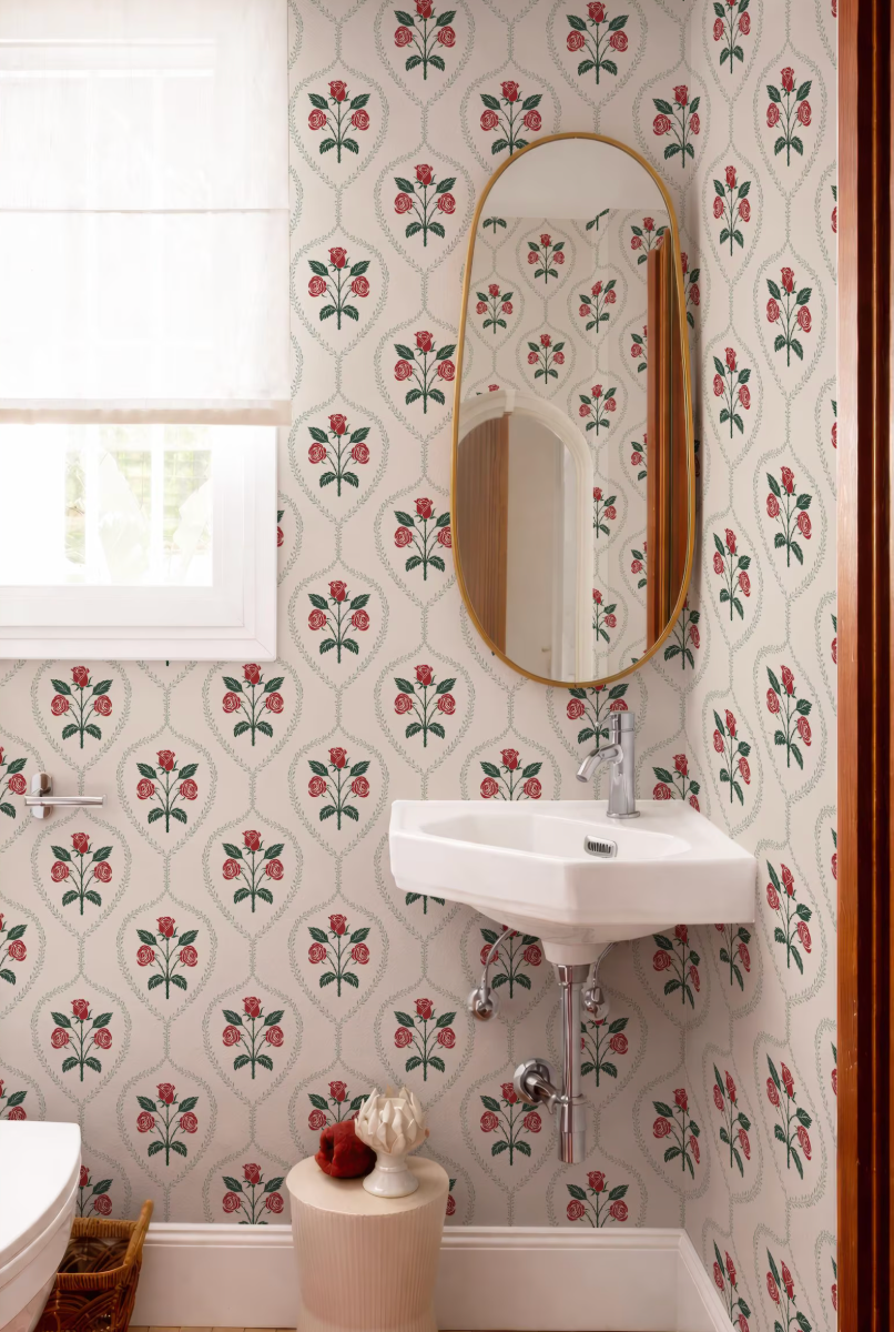 a bathroom with a white sink and a large mirror above it. The walls are covered in a floral patterned wallpaper, and there is a window in the background.