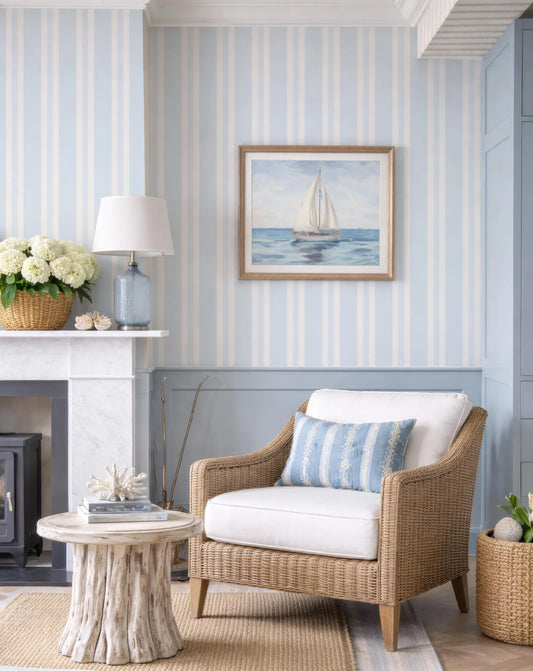 a cozy living room with a wicker chair, a white lamp, a framed painting of a sailboat, and a fireplace. The room has blue and white striped wallpaper, and the furniture and decor create a harmonious and inviting atmosphere.