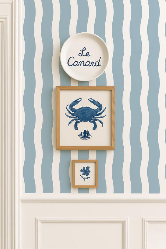 a wall with a blue and white striped wallpaper, and there are three framed pictures hanging on it. The top picture is a plate with the word "Le Camard" written on it, and the two other pictures are smaller framed images depicting a crab and a flower.