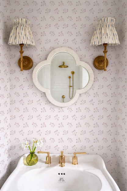 a bathroom with a white sink, a mirror, and two wall sconces. The walls are covered in a floral pattern, and there is a vase of flowers on the sink.