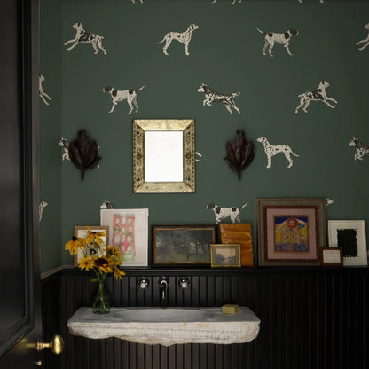 a bathroom with a green wall adorned with a pattern of white and black spotted dogs. The bathroom has a sink, a mirror, and various framed pictures and artwork displayed on the wall.