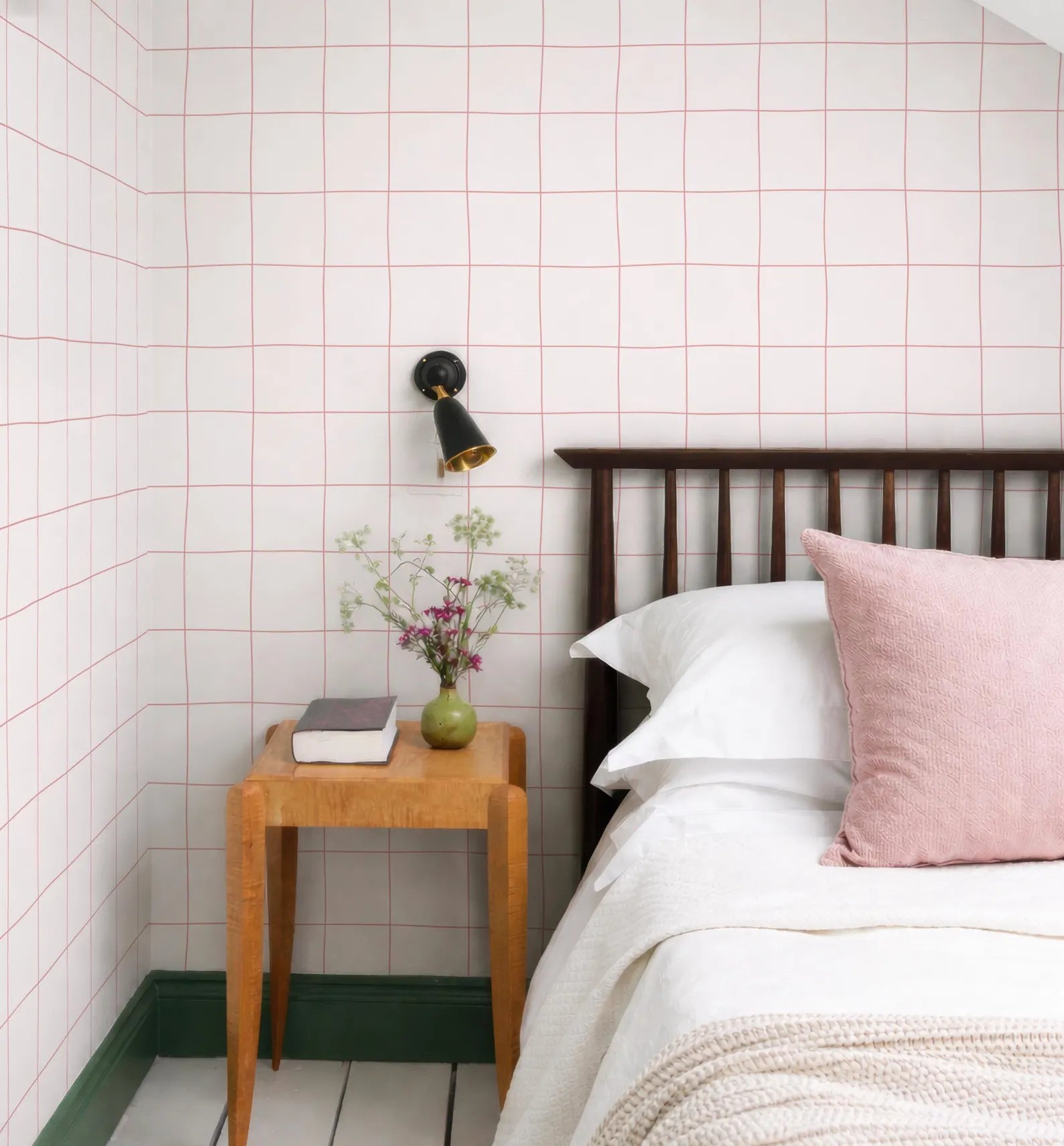 a cozy bedroom scene with a bed, a wooden nightstand, and a vase of flowers. The walls are covered in pink and white checkered wallpaper, and the room has a warm, inviting atmosphere.
