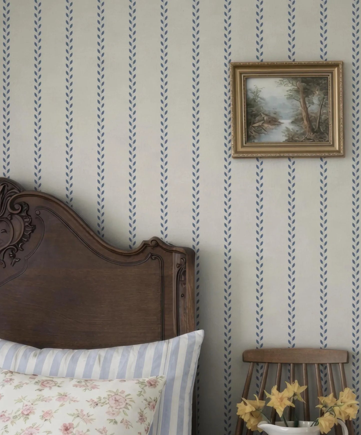 a bedroom with a wooden headboard, a framed landscape painting on the wall, and a wooden chair with yellow flowers on it.