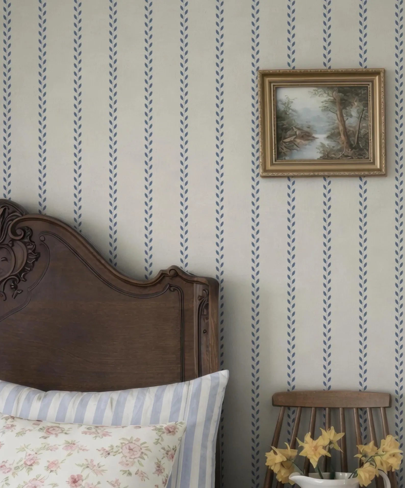 a bedroom with a wooden headboard, a framed landscape painting on the wall, and a wooden chair with yellow flowers on it.