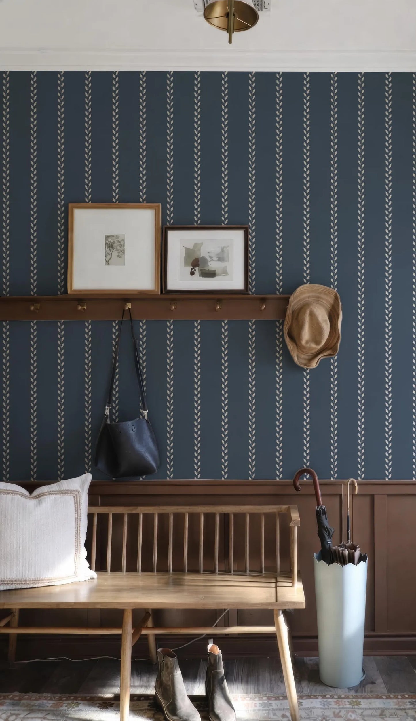 a cozy entryway with a wooden bench, a hat rack, and a pair of boots. The wall behind the bench has a blue and white striped pattern, and there are two framed pictures hanging on the wall. A wooden shelf is also present, holding a handbag and an umbrella.