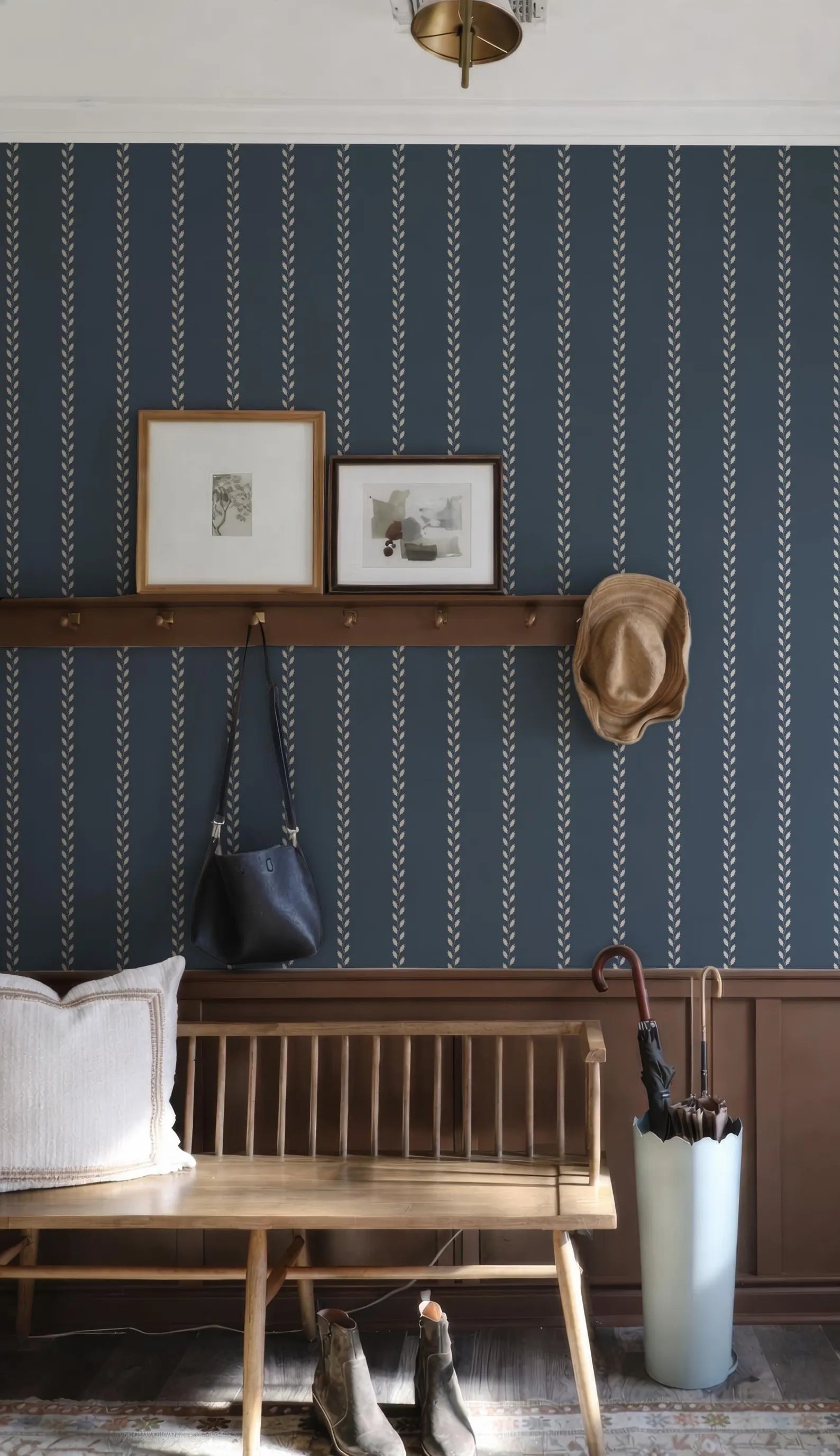 a cozy entryway with a wooden bench, a hat rack, and a pair of boots. The wall behind the bench has a blue and white striped pattern, and there are two framed pictures hanging on the wall. A wooden shelf is also present, holding a handbag and an umbrella.