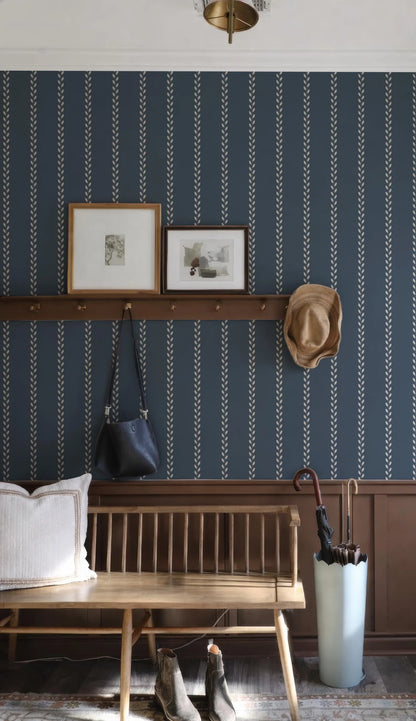 a cozy entryway with a wooden bench, a hat rack, and a pair of boots. The wall behind the bench has a blue and white striped pattern, and there are two framed pictures hanging on the wall. A wooden shelf is also present, holding a handbag and an umbrella.