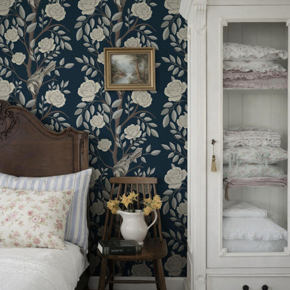 a bedroom with a wooden headboard, a blue floral wallpaper, and a white wooden cabinet with a glass door. The room also features a bed with a striped pillow, a wooden chair with a vase of yellow flowers, and a framed picture on the wall.