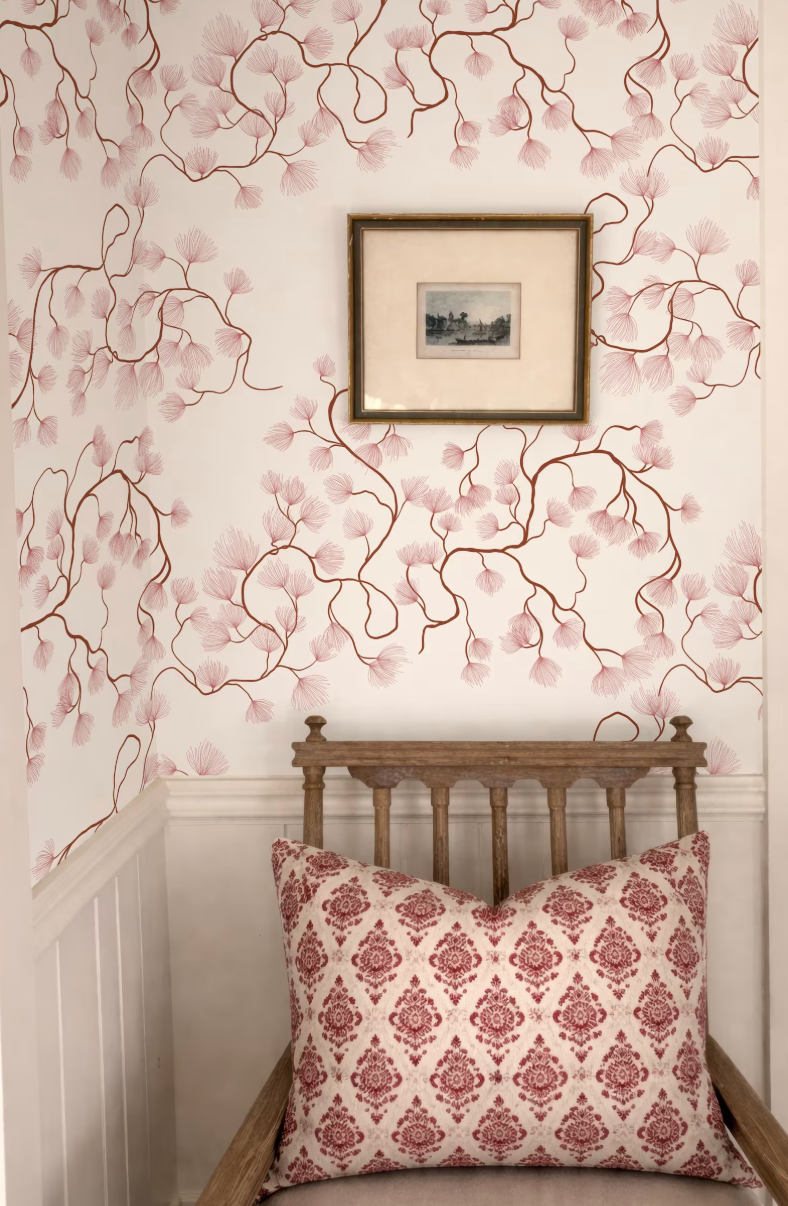 a wooden chair with a red and white patterned pillow, set against a wall with a floral wallpaper. Above the chair, there is a framed picture hanging on the wall.