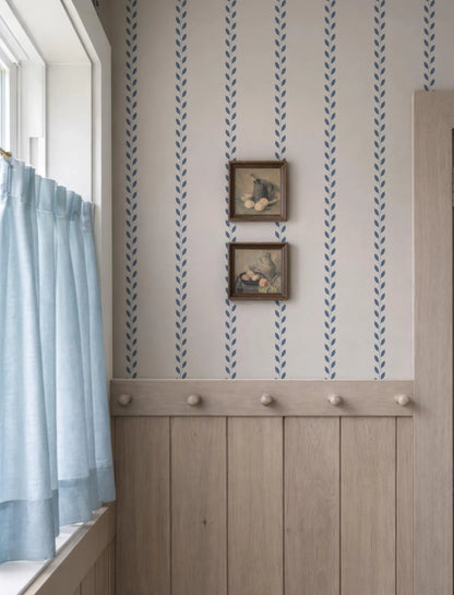 a room with a window, a wooden wall, and a wallpapered wall with a pattern of blue and white stripes. There are two framed pictures hanging on the wall, and a curtain is visible on the left side of the window.