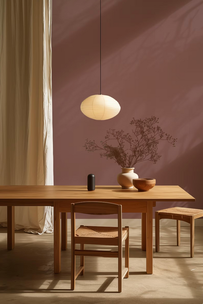 a minimalist dining room with a wooden table, two chairs, and a hanging light fixture. The table is set with a vase of dried flowers and a bowl, creating a serene and elegant atmosphere.