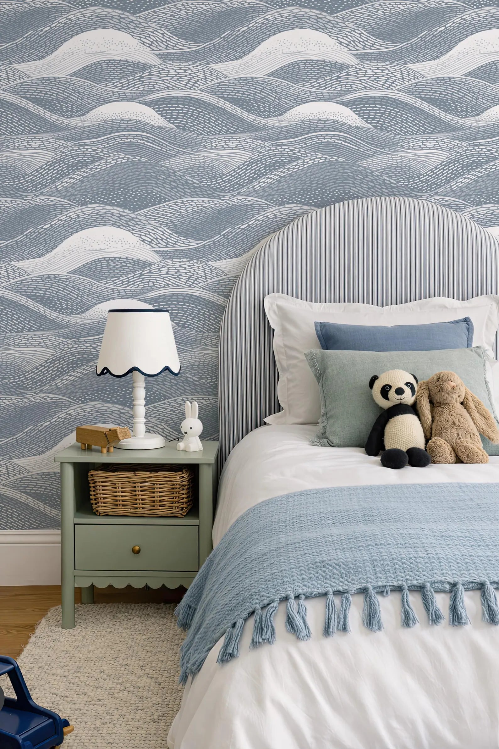 a cozy bedroom scene with a bed, nightstand, and stuffed animals. The bed has a blue and white striped headboard and a blue blanket. The nightstand has a white lamp and a small basket. There are two stuffed animals, a panda and a bunny, sitting on the bed. The walls are covered in a blue and white wallpaper with a wave pattern.
