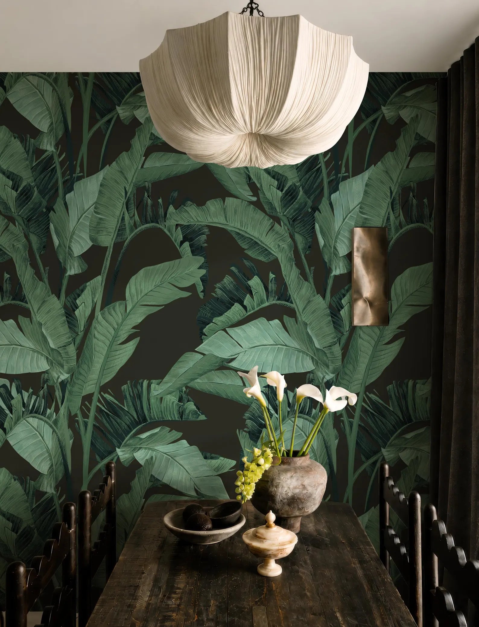 a dining room with a large green leafy wallpaper, a wooden table with a vase of flowers, and a chandelier hanging above.