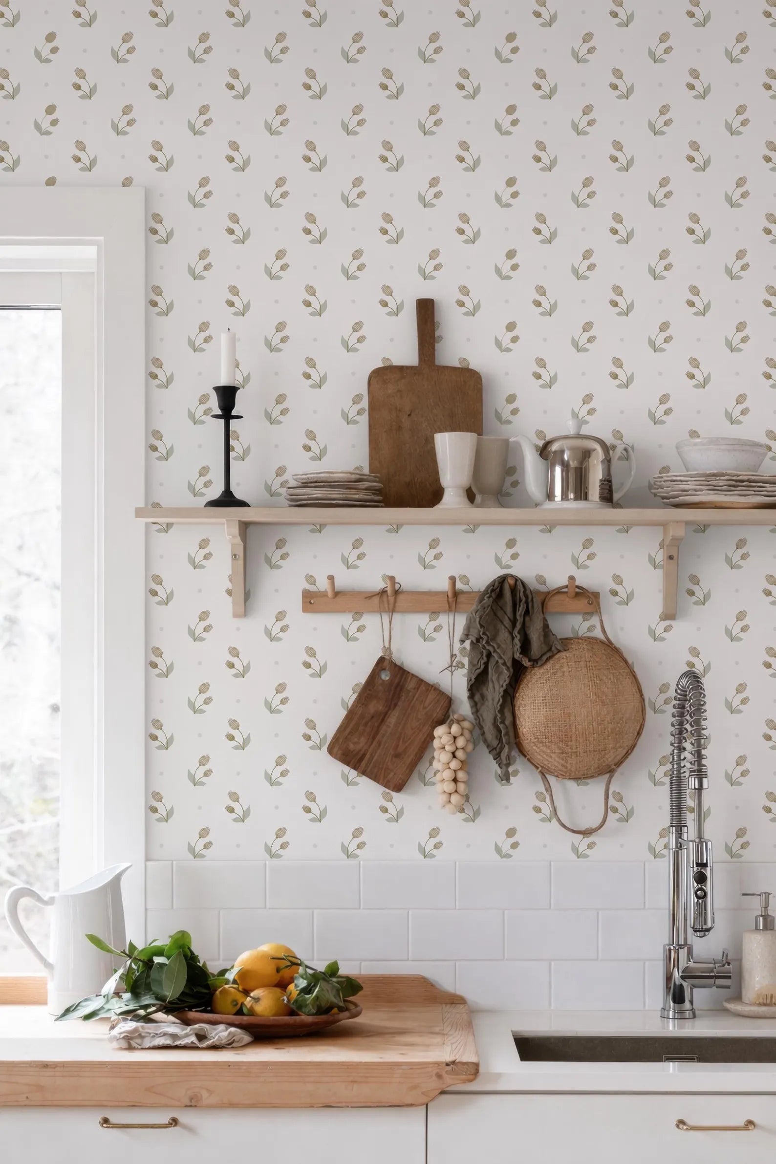 a kitchen with a white countertop, a wooden cutting board, a shelf with various kitchen items, and a sink. The wall behind the countertop has a patterned wallpaper with a repeating design of leaves and flowers.
