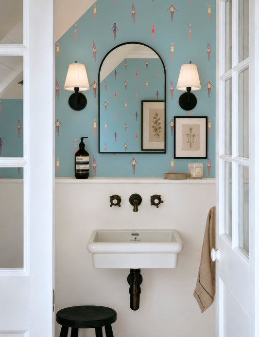 a bathroom with a white sink, a mirror, and a stool. The walls are decorated with a patterned wallpaper featuring small figures. There are two light fixtures above the mirror, and a towel is hanging on the wall.