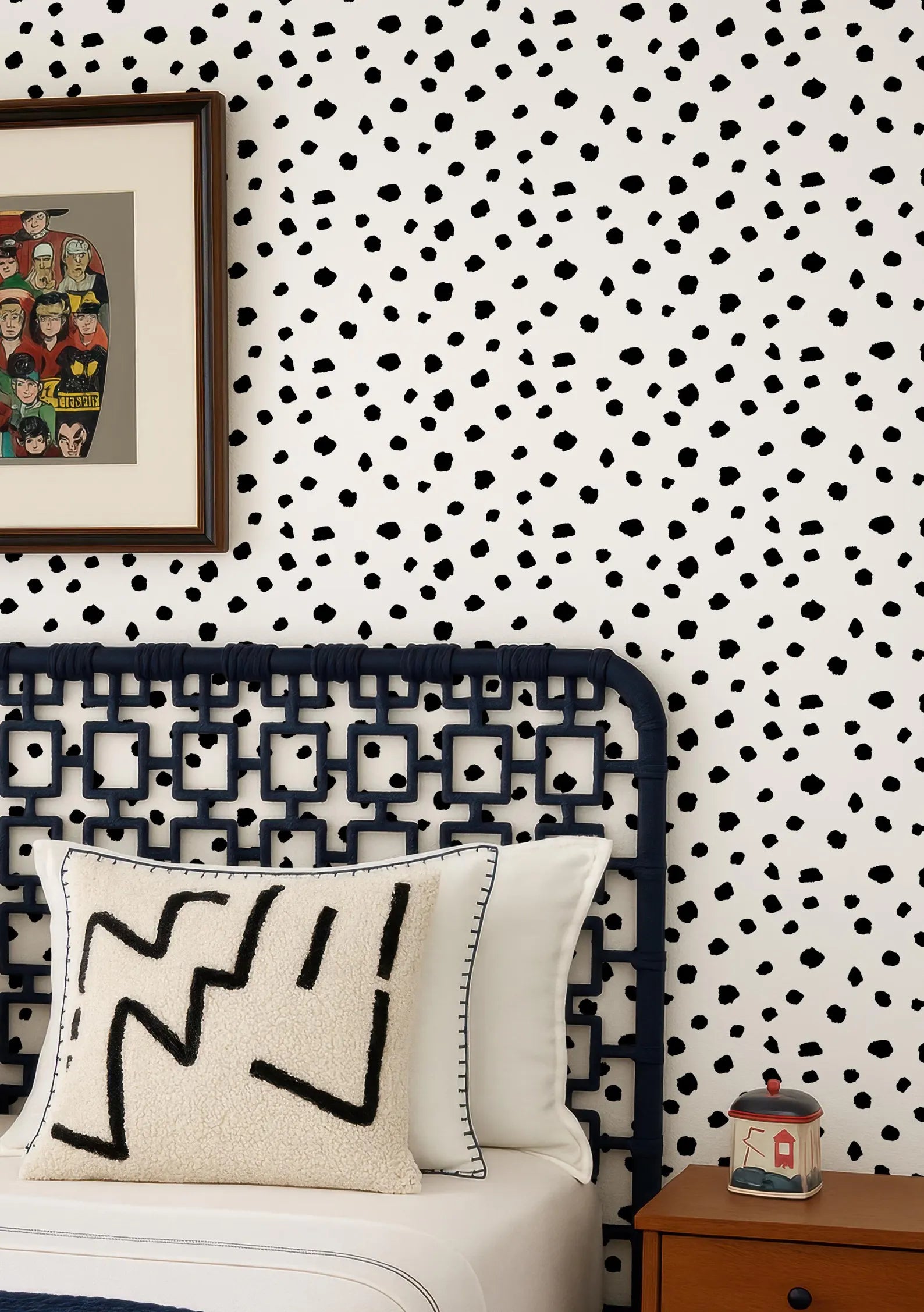 a bedroom with a black metal headboard, a white pillow with a black geometric pattern, and a wooden nightstand with a small container on it. The wall behind the bed has a black and white polka dot pattern.
