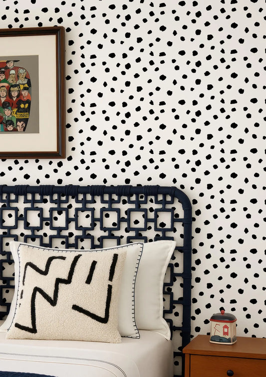 a bedroom with a black metal headboard, a white pillow with a black geometric pattern, and a wooden nightstand with a small container on it. The wall behind the bed has a black and white polka dot pattern.
