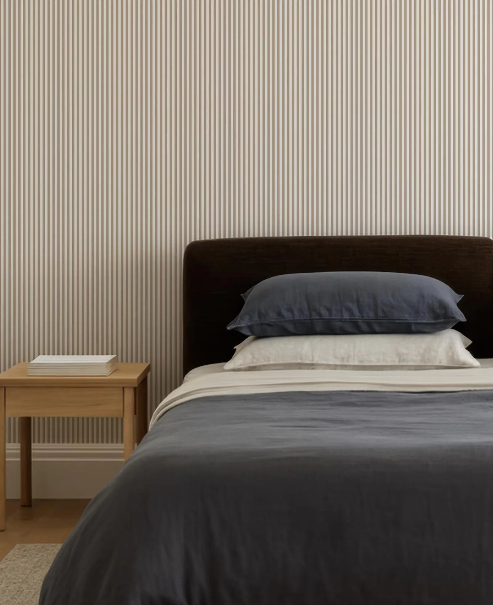 a neatly made bed with a dark brown headboard and a gray comforter. The bed is set against a striped wallpaper, creating a cozy and inviting atmosphere.