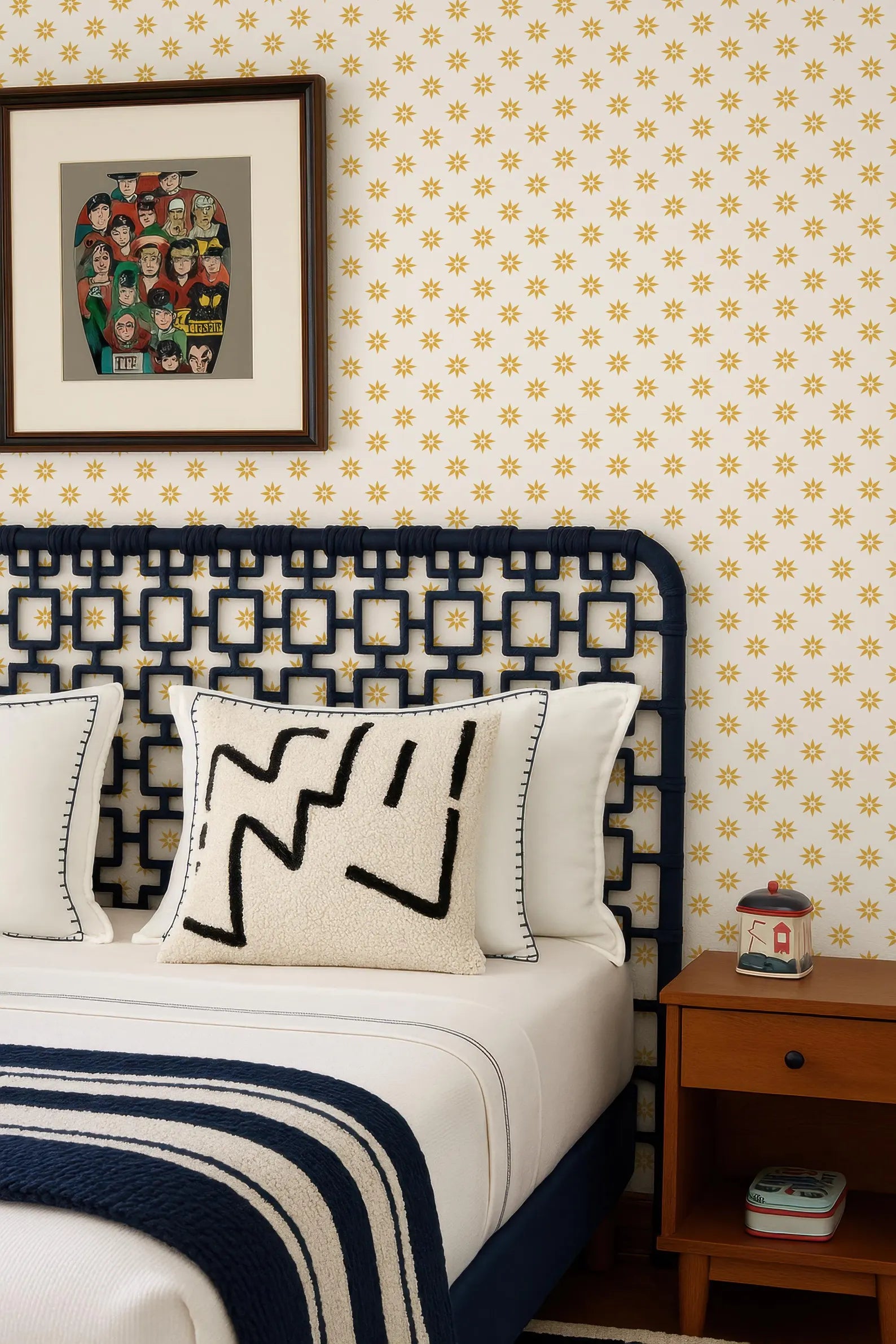 a bedroom with a bed, a nightstand, and a framed picture on the wall. The bed has a black headboard and white pillows, and the wall is covered in yellow and white patterned wallpaper.