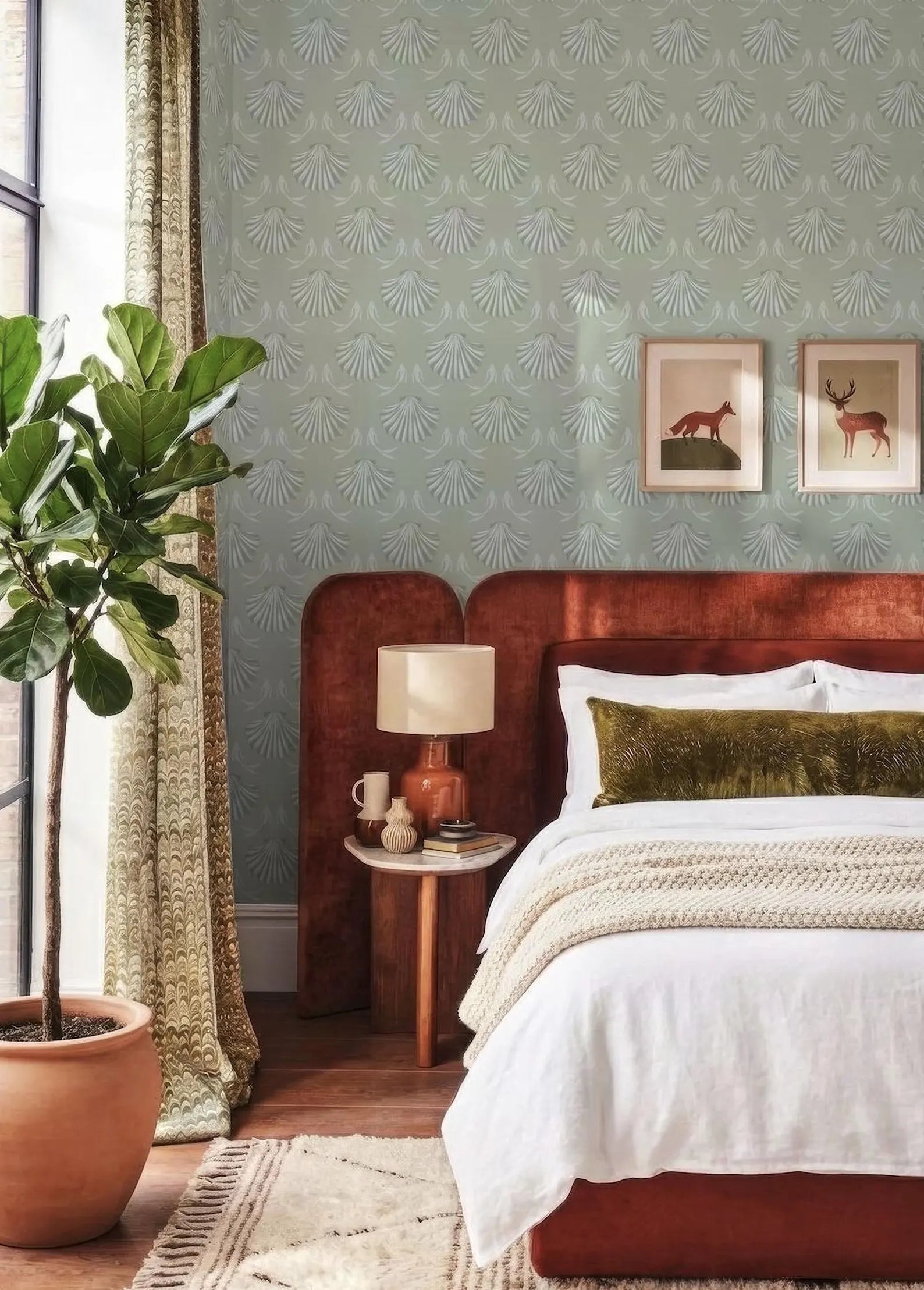 a cozy bedroom with a large bed, a wooden nightstand, and a potted plant. The walls are covered in a patterned wallpaper, and there are two framed pictures on the wall. The room has a warm and inviting atmosphere, with a mix of natural and modern elements.