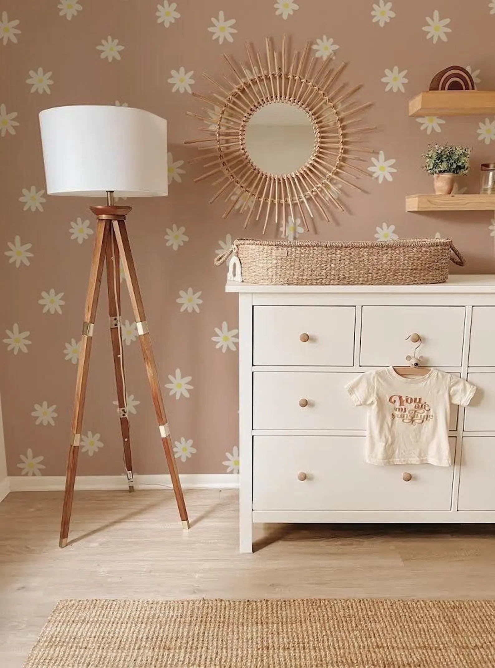 a nursery room with a white dresser, a tripod lamp, a mirror, and a basket. The walls are decorated with a floral pattern, and there is a baby changing pad in the room.