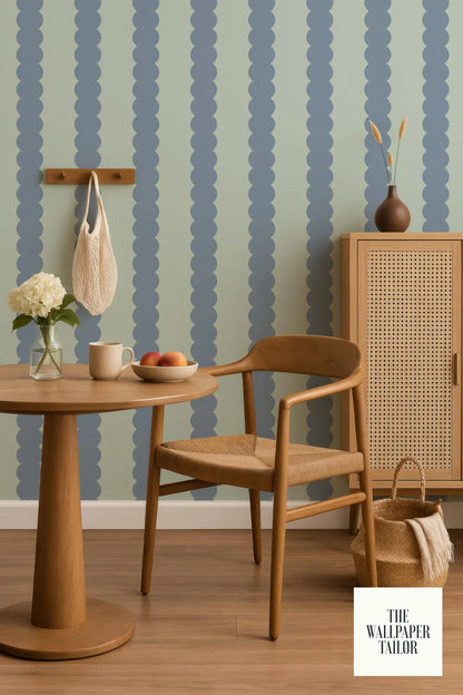 a cozy corner of a room with a wooden chair, a round wooden table, and a woven basket. The walls are adorned with a pattern of blue and green stripes, and there are some decorative elements such as a vase with flowers and a bowl of fruit on the table.