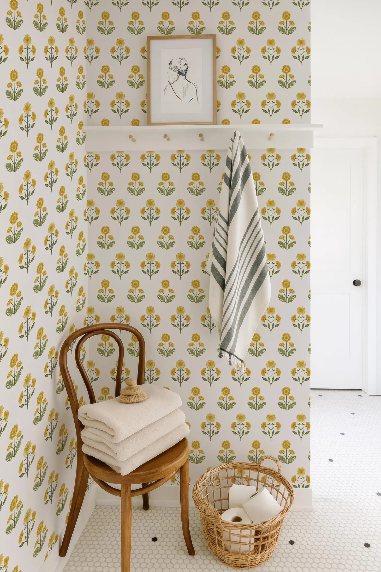 a bathroom corner with a wooden chair, a towel rack with a striped towel, and a basket of toilet paper. The walls are covered in a floral wallpaper pattern, and there is a framed picture hanging above the chair.