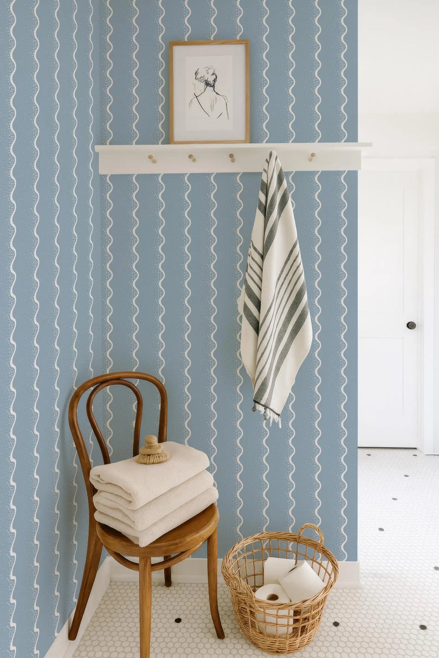 a bathroom with a blue and white striped wallpaper, a wooden chair with folded towels on it, a basket of toilet paper, and a framed picture on a shelf above the chair.