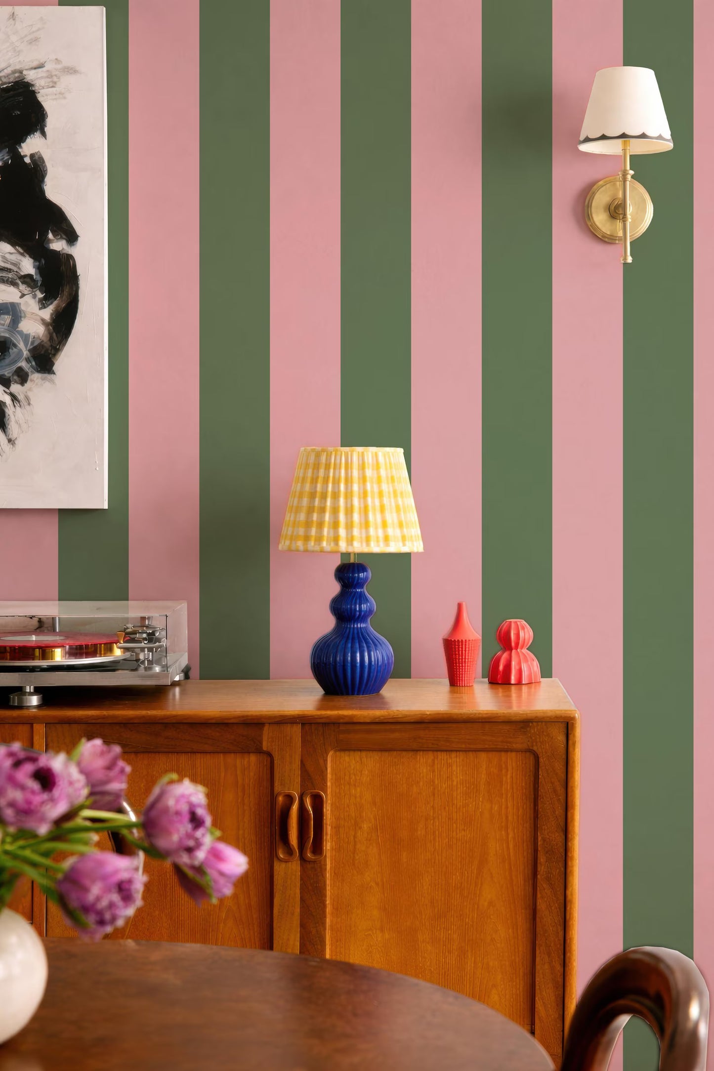 a vibrant and eclectic interior design with a mix of pink, green, and blue stripes on the walls. A wooden cabinet with a blue lamp and a red vase on top is situated in the foreground, while a framed artwork hangs on the wall behind it. A white lamp is mounted on the wall, and a vase of purple flowers is placed on a table in the foreground.