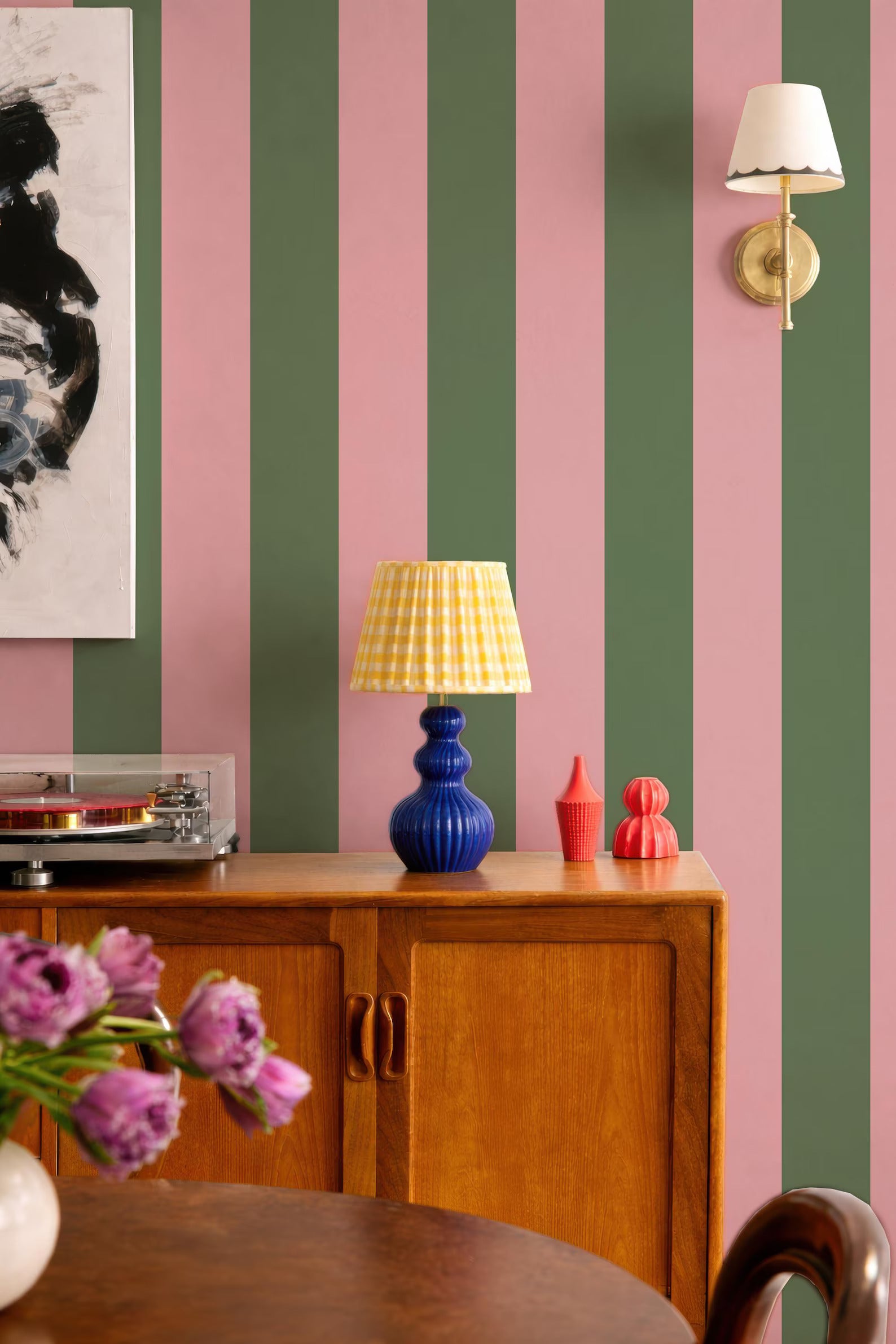 a vibrant and eclectic interior design with a mix of pink, green, and blue stripes on the walls. A wooden cabinet with a blue lamp and a red vase on top is situated in the foreground, while a framed artwork hangs on the wall behind it. A white lamp is mounted on the wall, and a vase of purple flowers is placed on a table in the foreground.