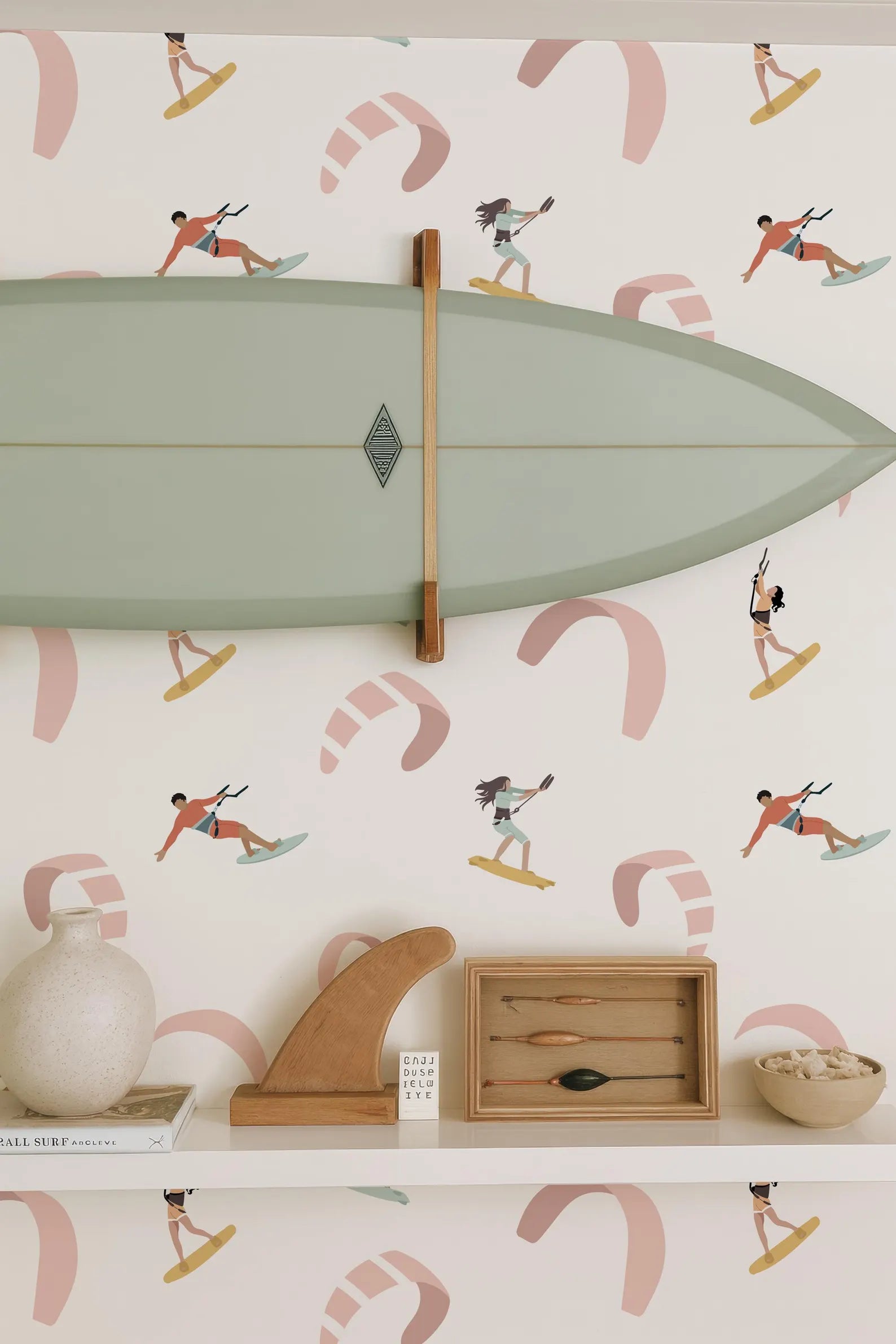 a wall with a surfboard mounted on it, along with various decorative items such as a wooden surfboard holder, a wooden sculpture, and a framed picture of a surfer. The wall also has a pattern of surfers and kites, creating a beach-themed atmosphere.