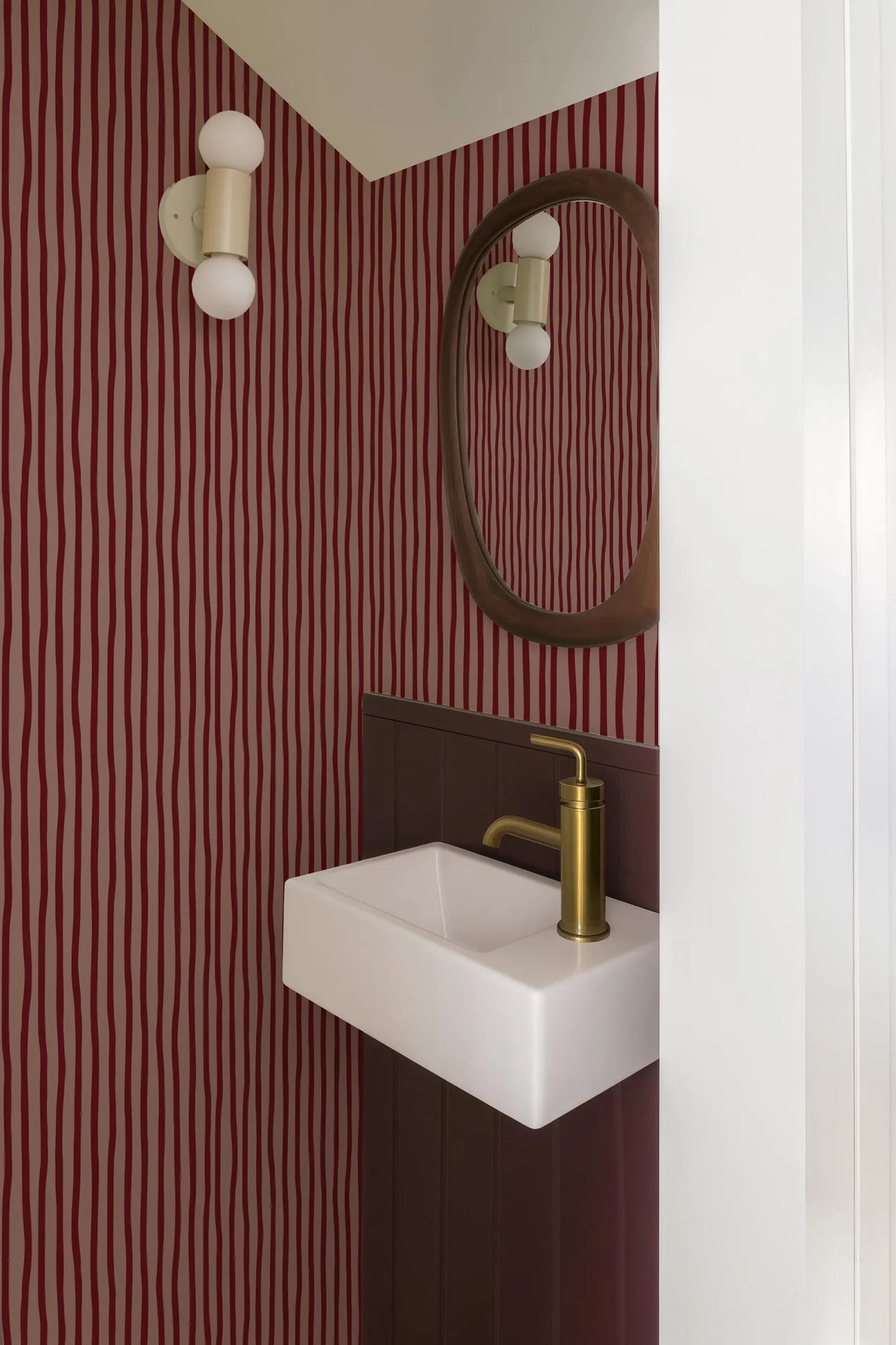 a bathroom with a white sink and a gold faucet, set against a red and white striped wallpaper. There is a mirror above the sink, and the walls are painted in a maroon color.