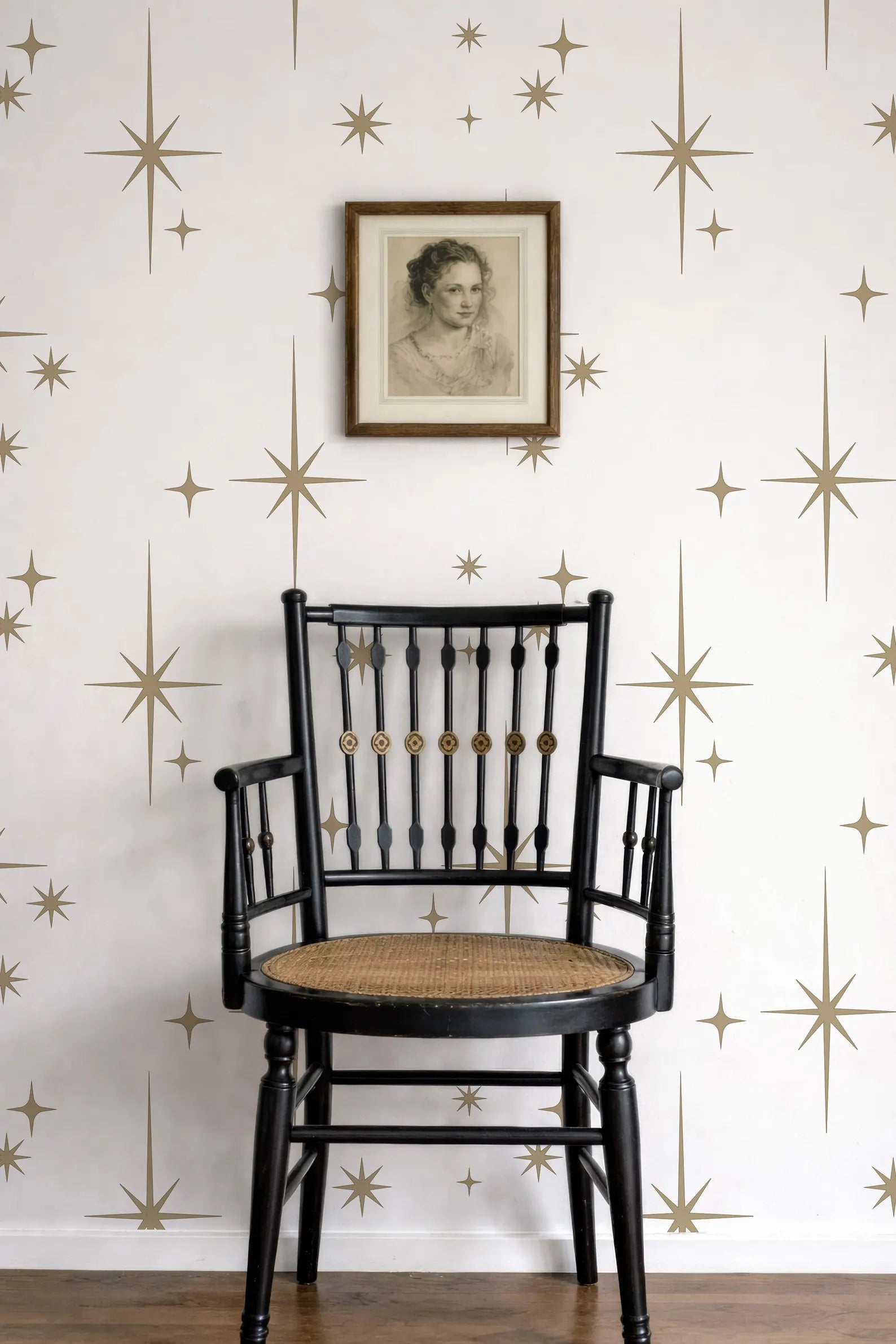 a black wooden chair with a woven seat, positioned against a white wall with gold star patterns. Above the chair, there is a framed portrait of a woman.