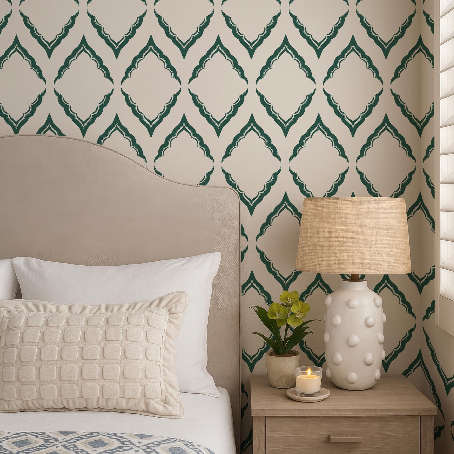 a cozy bedroom scene with a bed, a nightstand, and a lamp. The bed has a beige headboard and white pillows, and the nightstand has a white lamp with a beige shade. The wall behind the bed has a patterned wallpaper with green and white diamond shapes.