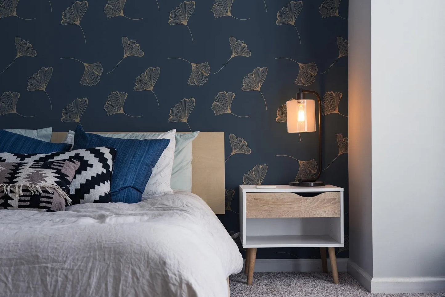 a cozy bedroom with a bed adorned with white linens and colorful throw pillows, complemented by a wooden nightstand with a lamp and a white drawer. The wall behind the bed is decorated with a pattern of gold leaf-like designs in shades of blue and white, creating a serene and inviting atmosphere.