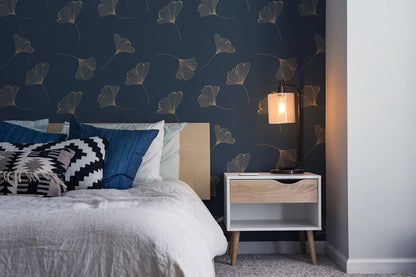a cozy bedroom with a bed adorned with white linens and colorful throw pillows, complemented by a wooden nightstand with a lamp and a white drawer. The wall behind the bed is decorated with a pattern of gold leaf-like designs in shades of blue and white, creating a serene and inviting atmosphere.