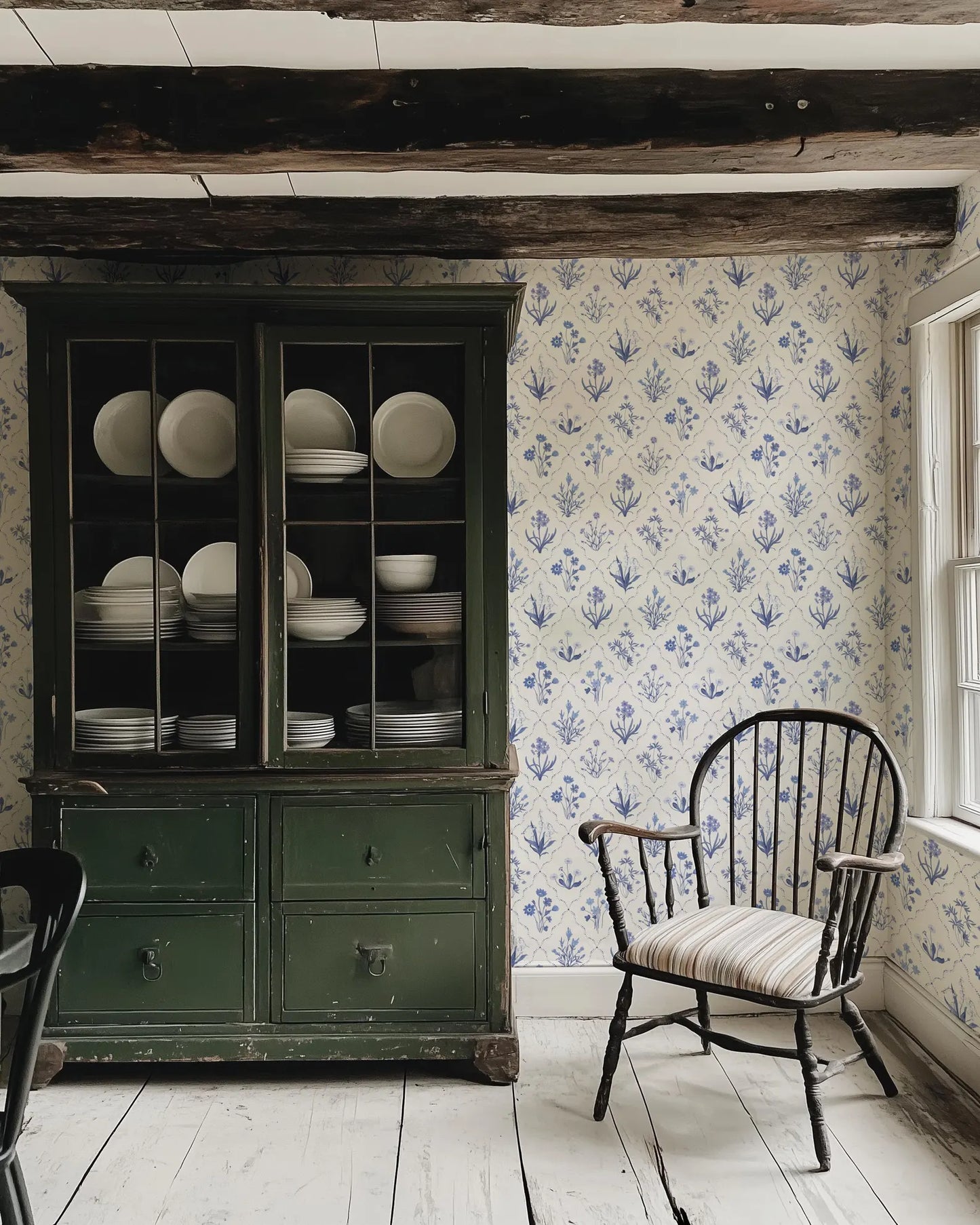 a vintage green china cabinet with a glass door, displaying an array of white plates and bowls. A black wooden chair is positioned in front of the cabinet, and the walls are adorned with a blue and white floral wallpaper.
